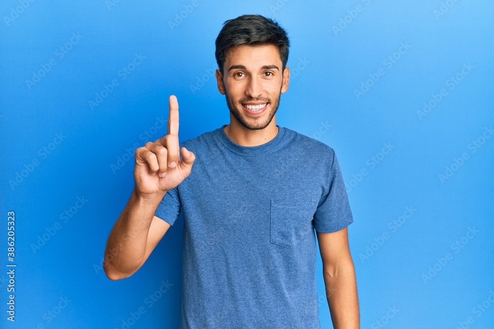 Young handsome man wearing casual tshirt over blue background smiling with an idea or question pointing finger up with happy face, number one Young handsome man wearing casual tshirt over blue background smiling with an idea or question pointing finger up with happy face, number one