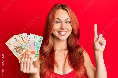 Young beautiful redhead woman holding norwegian krone banknotes surprised with an idea or question pointing finger with happy face, number one photo