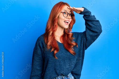 Young beautiful redhead woman wearing casual sweater and glasses over blue background very happy and smiling looking far away with hand over head. searching concept.