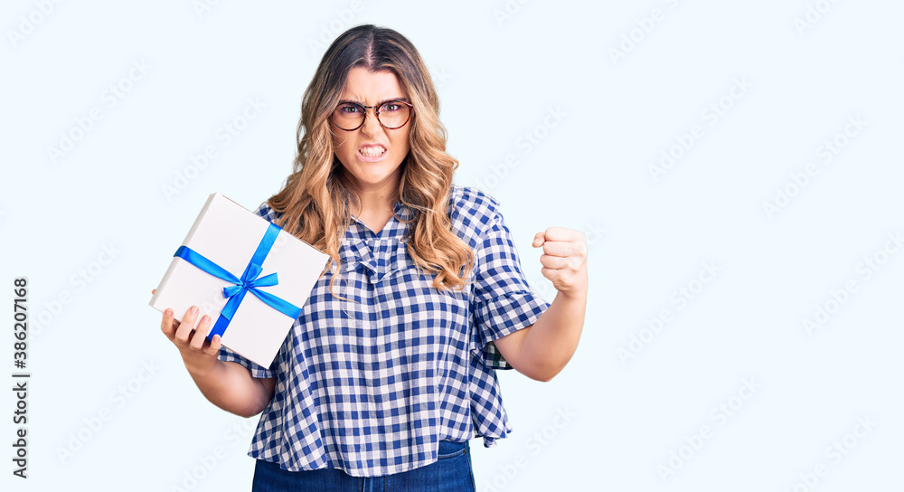 Young caucasian woman holding gift annoyed and frustrated shouting with anger, yelling crazy with anger and hand raised Young caucasian woman holding gift annoyed and frustrated shouting with anger, yelling crazy with anger and hand raised