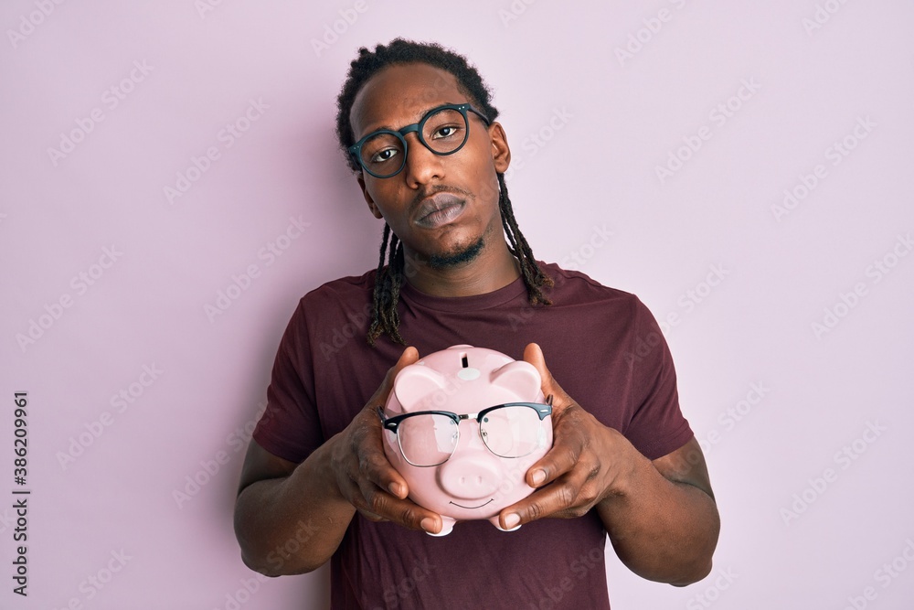 African american man with braids holding piggy bank with glasses relaxed with serious expression on face. simple and natural looking at the camera. African american man with braids holding piggy bank with glasses relaxed with serious expression on face. simple and natural looking at the camera.
