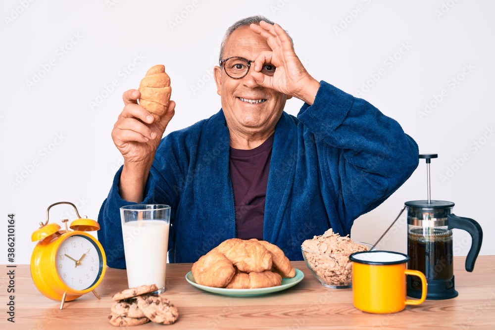 Senior handsome man with gray hair sitting on the table eating croissant for breakfast smiling happy doing ok sign with hand on eye looking through fingers Senior handsome man with gray hair sitting on the table eating croissant for breakfast smiling happy doing ok sign with hand on eye looking through fingers