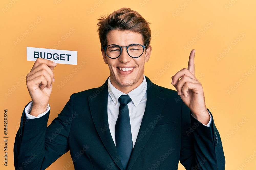 Handsome caucasian business man holding budget word on paper surprised with an idea or question pointing finger with happy face, number one Handsome caucasian business man holding budget word on paper surprised with an idea or question pointing finger with happy face, number one