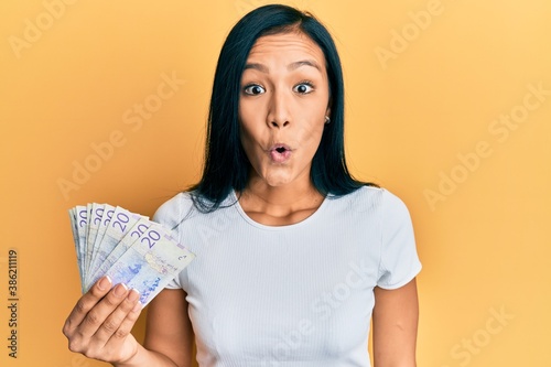 Beautiful hispanic woman holding 20 swedish krona banknotes scared and amazed with open mouth for surprise, disbelief face photo