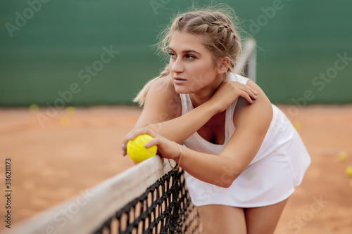 Relaxing after tennis training. Young beautiful girl in white uniform and sporty cap on a tennis court near the net © volody10