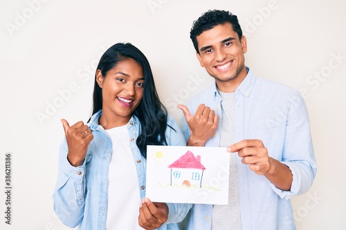 Beautiful latin young couple showing house proyect on a paper pointing thumb up to the side smiling happy with open mouth photo