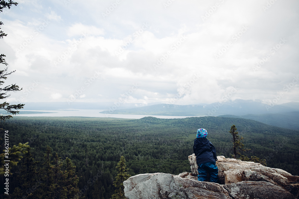 a child stands on a mountain and looks into the distance, children's Hiking trip, kid high in the mountains, family tourism