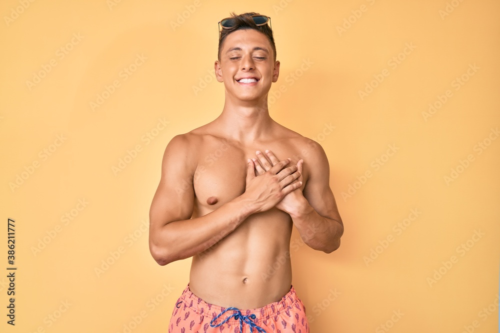 Fototapeta premium Young hispanic boy wearing swimwear shirtless smiling with hands on chest with closed eyes and grateful gesture on face. health concept.