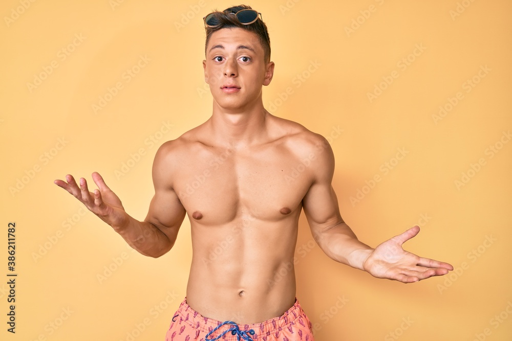 Young hispanic boy wearing swimwear shirtless clueless and confused expression with arms and hands raised. doubt concept.