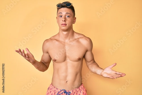 Young hispanic boy wearing swimwear shirtless clueless and confused expression with arms and hands raised. doubt concept.