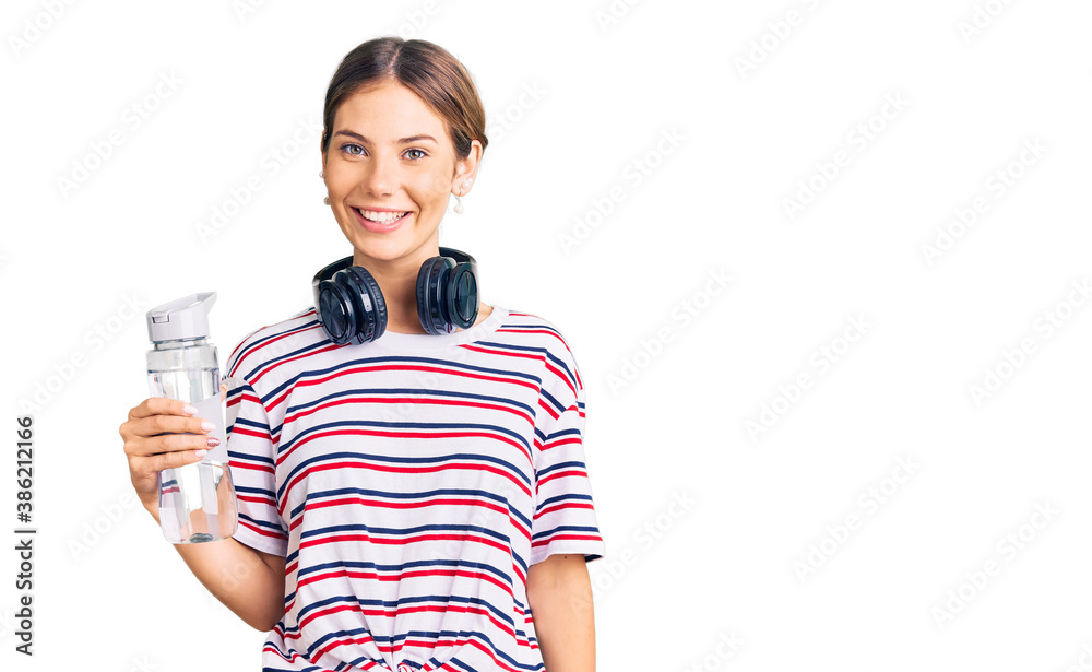 Beautiful caucasian woman with blonde hair wearing sportswear and headphones drinking a bottle of water looking positive and happy standing and smiling with a confident smile showing teeth Beautiful caucasian woman with blonde hair wearing sportswear and headphones drinking a bottle of water looking positive and happy standing and smiling with a confident smile showing teeth
