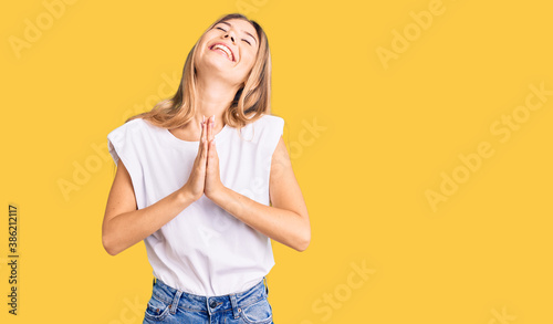 Beautiful caucasian woman with blonde hair wearing casual white tshirt begging and praying with hands together with hope expression on face very emotional and worried. begging.