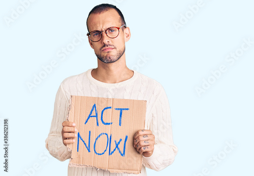 Young handsome man holding act now banner thinking attitude and sober expression looking self confident