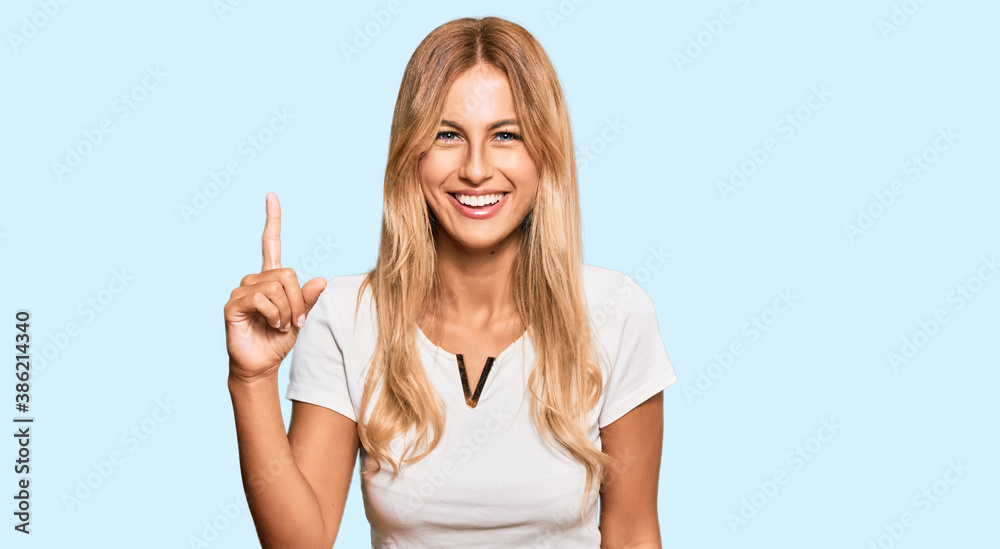 Beautiful blonde young woman wearing casual white tshirt pointing finger up with successful idea. exited and happy. number one.