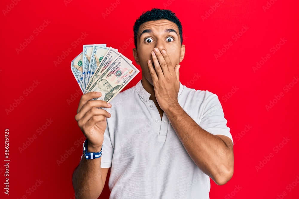 Young arab man holding dollars covering mouth with hand, shocked and afraid for mistake. surprised expression Young arab man holding dollars covering mouth with hand, shocked and afraid for mistake. surprised expression