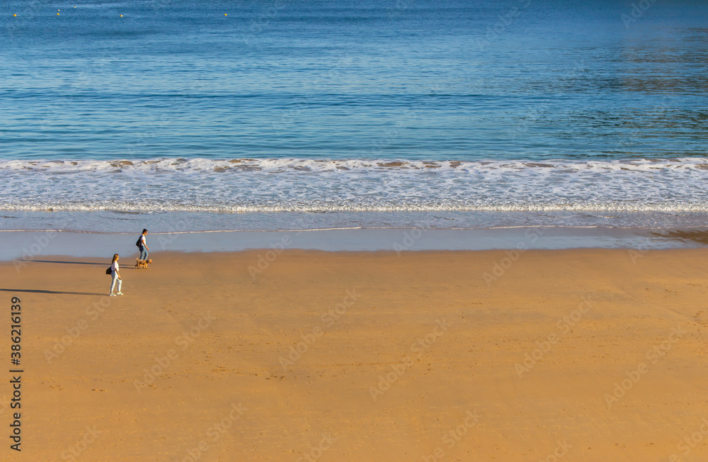 San Sebastian, Spain - 10/11/2019: Unknown people with dog walking on the beach. Tourists in bay of Biscay in San Sebastian in autumn. Wide aerial beach in morning sunlight. Active people. San Sebastian, Spain - 10/11/2019: Unknown people with dog walking on the beach. Tourists in bay of Biscay in San Sebastian in autumn. Wide aerial beach in morning sunlight. Active people.