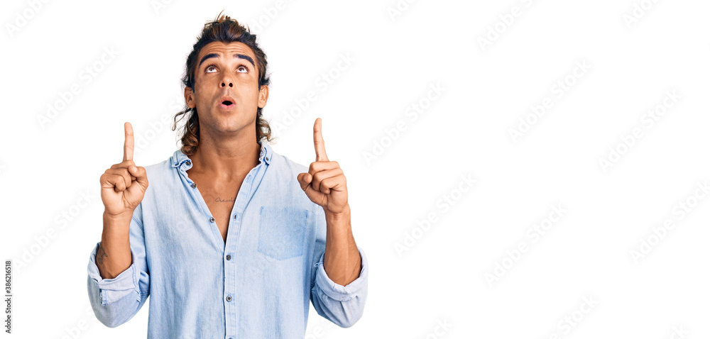 Young hispanic man wearing summer style amazed and surprised looking up and pointing with fingers and raised arms.