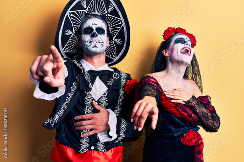 Young couple wearing mexican day of the dead costume over yellow laughing at you, pointing finger to the camera with hand over body, shame expression