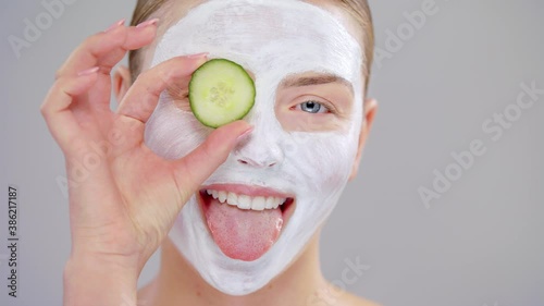 Extreme Closeup Portrait Young Caucasian Beautiful Blonde Woman With Blue Eyes She Covering Eye Fresh Cucumber White Mask Cosmetic On Face And Smiling On Gray Background Skin Care Concept.Slow Motion.