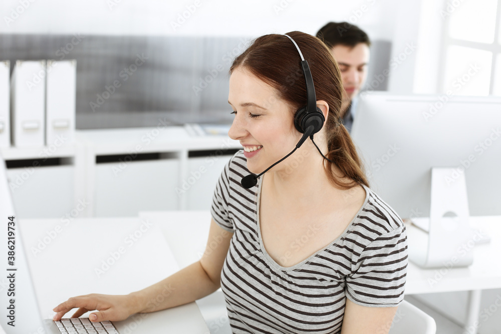 Naklejka premium Casual dressed young woman using headset and computer while talking with customers online. Group of operators at work. Call center, business concept