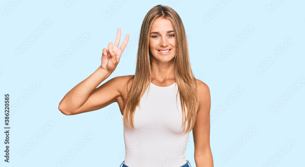 Young blonde woman wearing casual style with sleeveless shirt smiling looking to the camera showing fingers doing victory sign. number two.