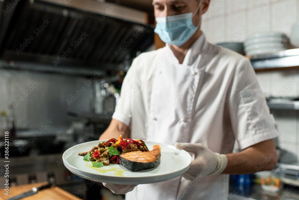 Young professional chef holding plate with fried salmon and vegetable garnish