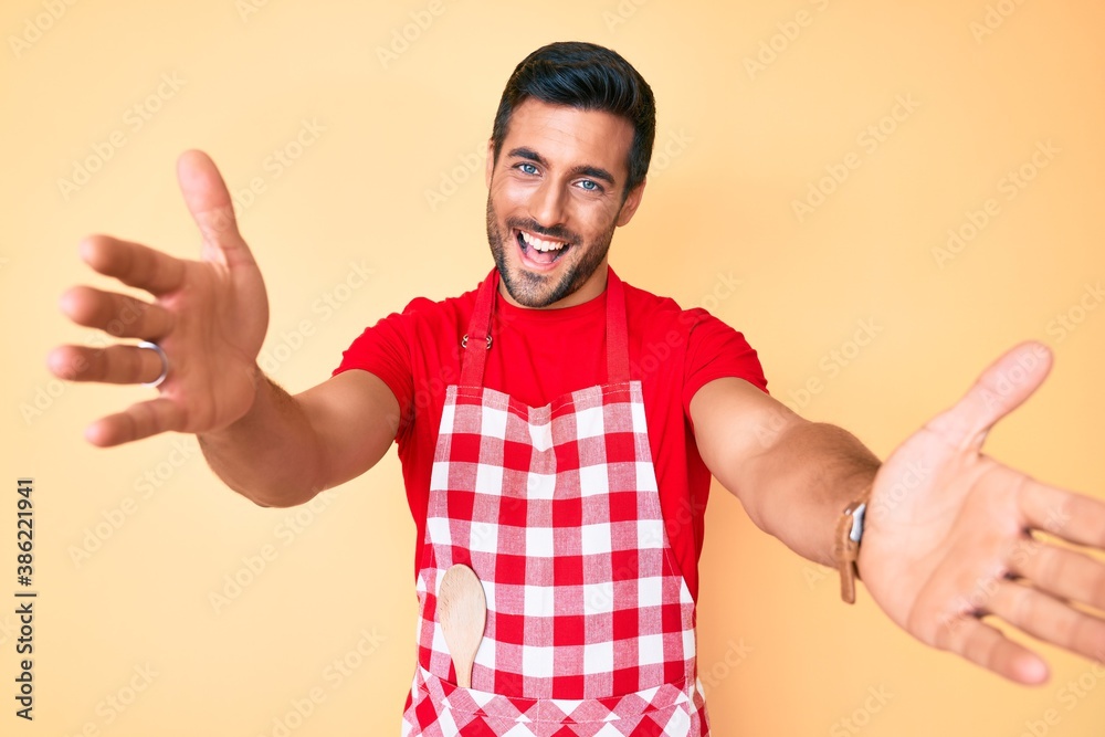 Young hispanic man wearing professional baker apron looking at the camera smiling with open arms for hug. cheerful expression embracing happiness. Young hispanic man wearing professional baker apron looking at the camera smiling with open arms for hug. cheerful expression embracing happiness.