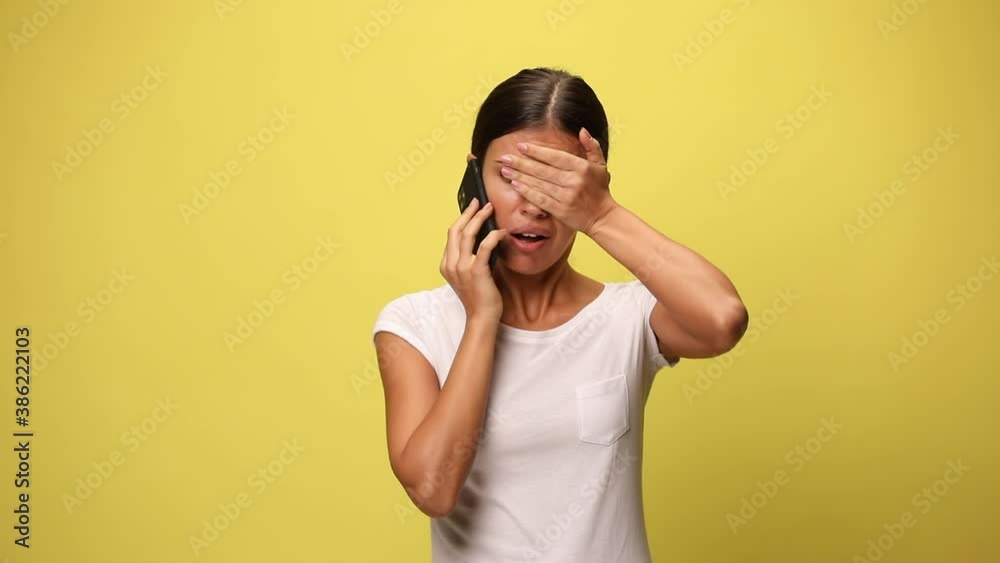 attractive casual woman biting her nails, talking on the phone, feeling anxious, slapping her face and gesturing a mad sign on yellow background attractive casual woman biting her nails, talking on the phone, feeling anxious, slapping her face and gesturing a mad sign on yellow background