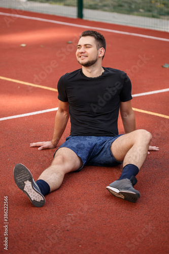 A young male athlete with a beard sits on a red tennis court. Handsome guy in t-shirt and shorts