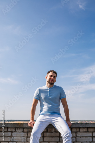 An attractive cool guy in a blue T-shirt with a beard sits against the background of the blue sky.