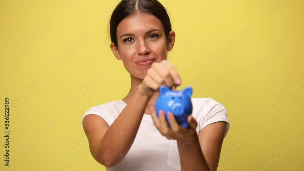 casual woman putting money in a piggy bank, aprroving, pointing at the camera on yellow background casual woman putting money in a piggy bank, aprroving, pointing at the camera on yellow background