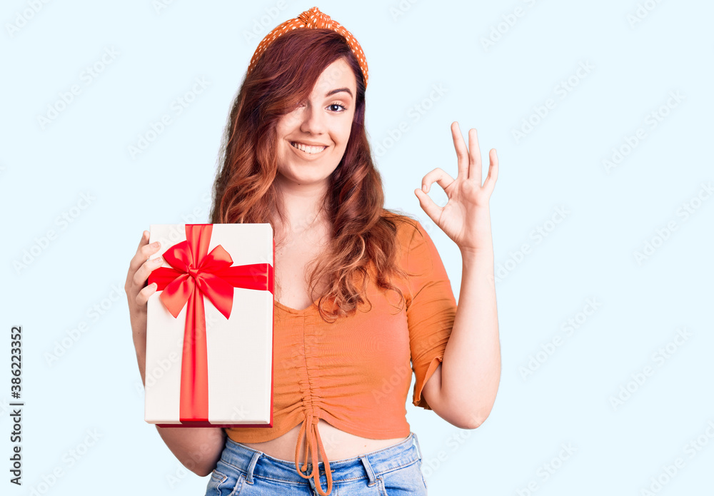Young beautiful woman holding gift doing ok sign with fingers, smiling friendly gesturing excellent symbol
