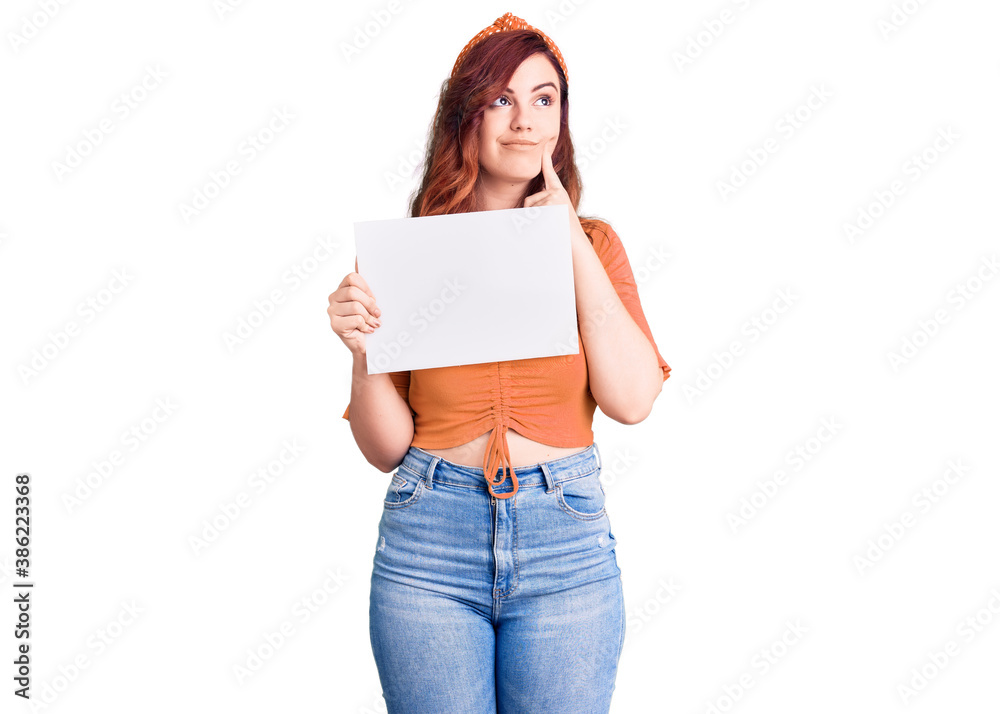 Young beautiful woman holding cardboard banner with blank space serious face thinking about question with hand on chin, thoughtful about confusing idea Young beautiful woman holding cardboard banner with blank space serious face thinking about question with hand on chin, thoughtful about confusing idea