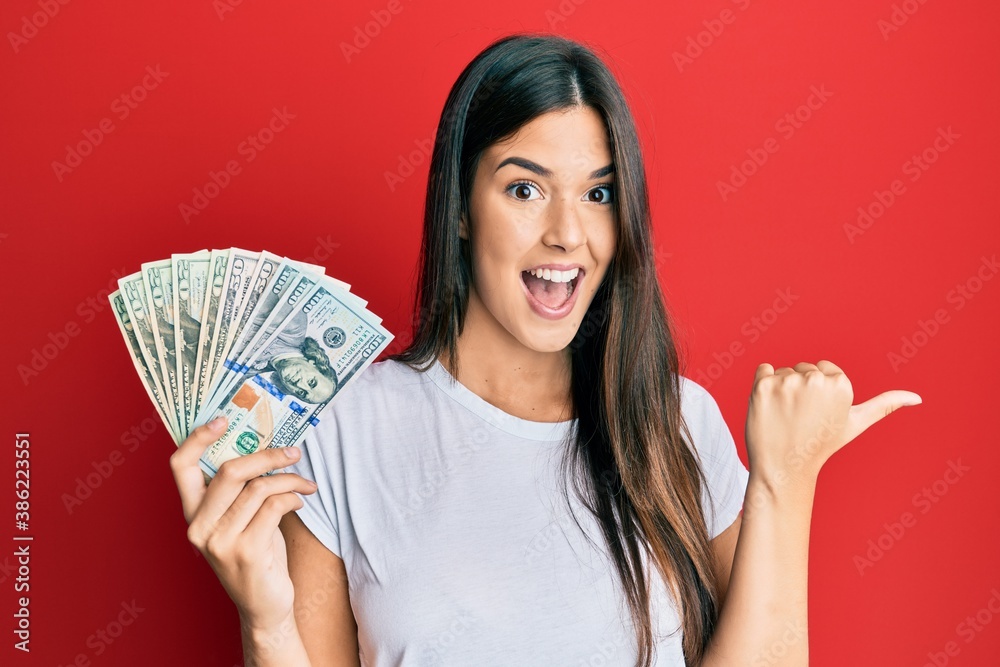 Young brunette woman holding dollars pointing thumb up to the side smiling happy with open mouth Young brunette woman holding dollars pointing thumb up to the side smiling happy with open mouth