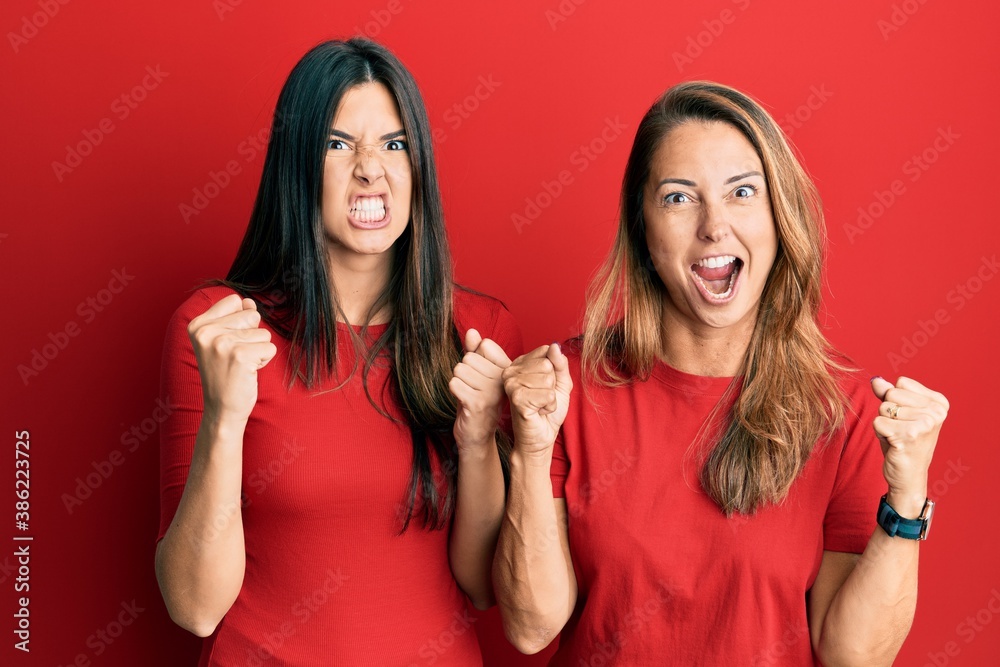 Hispanic family of mother and daughter wearing casual clothes over red background angry and mad raising fists frustrated and furious while shouting with anger. rage and aggressive concept. Hispanic family of mother and daughter wearing casual clothes over red background angry and mad raising fists frustrated and furious while shouting with anger. rage and aggressive concept.
