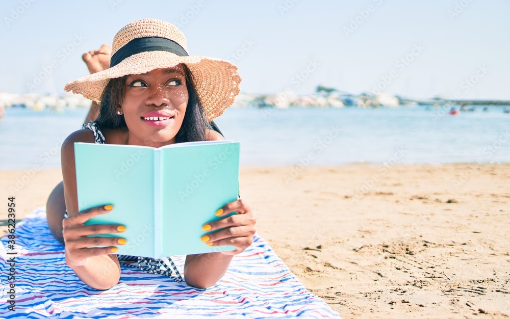 Young african american woman on vacation laying on the towel reading book at the beach