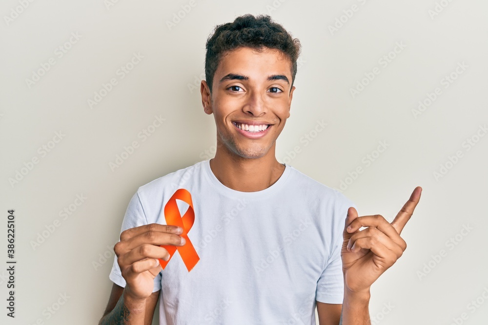 Young handsome african american man holding awareness orange ribbon smiling happy pointing with hand and finger to the side Young handsome african american man holding awareness orange ribbon smiling happy pointing with hand and finger to the side
