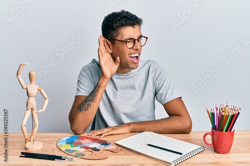 Young handsome african american man painter sitting palette and art manikin smiling with hand over ear listening an hearing to rumor or gossip. deafness concept.