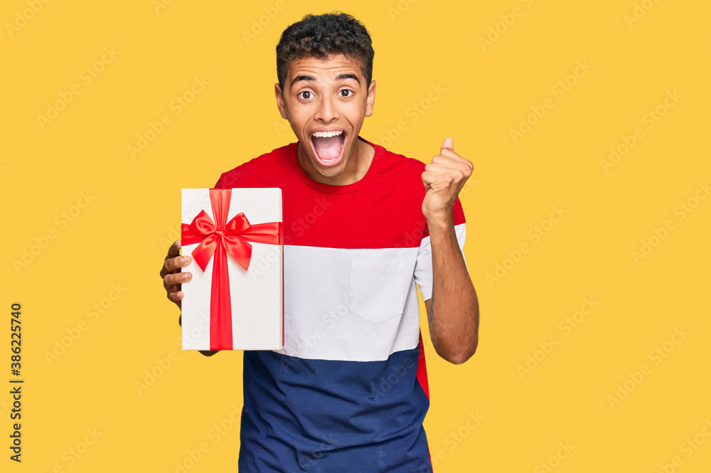 Young handsome african american man holding gift screaming proud, celebrating victory and success very excited with raised arms Young handsome african american man holding gift screaming proud, celebrating victory and success very excited with raised arms