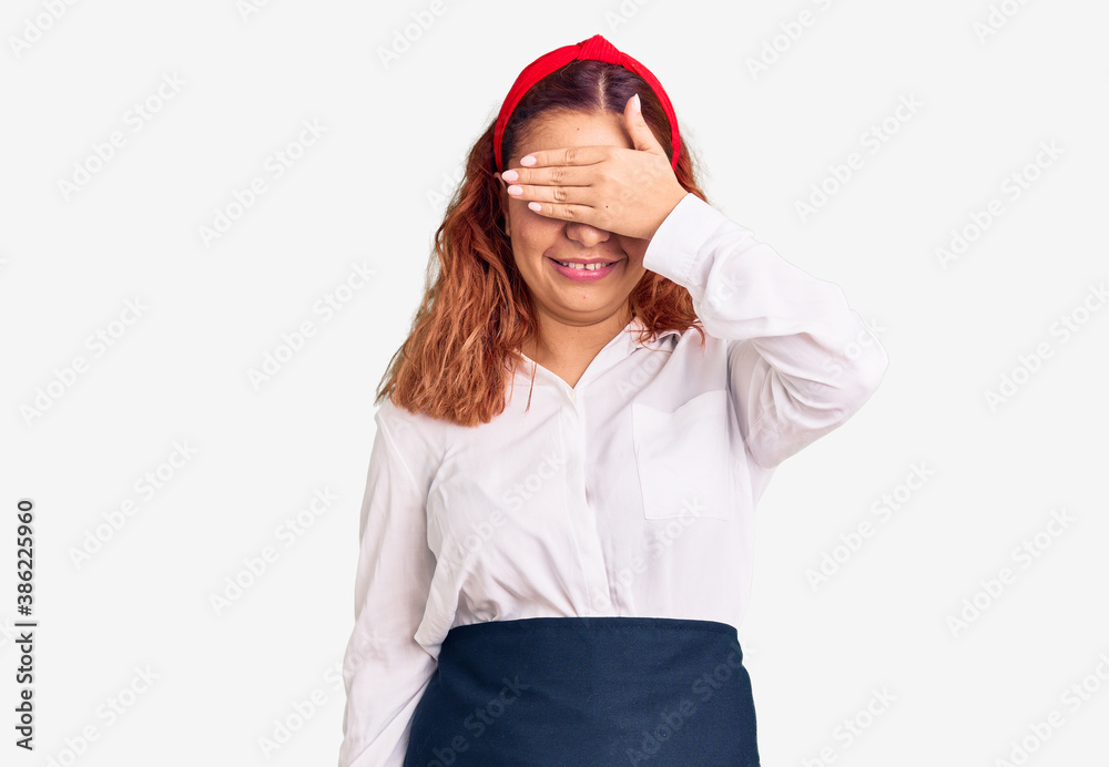 Young latin woman wearing waitress apron smiling and laughing with hand on face covering eyes for surprise. blind concept. Young latin woman wearing waitress apron smiling and laughing with hand on face covering eyes for surprise. blind concept.