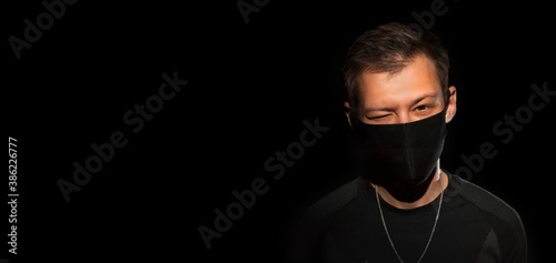 The guy in black clothes and a black medical mask on a black background winks and looks at the camera. Close-up. copy space