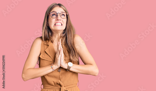 Young beautiful blonde woman wearing casual clothes and glasses begging and praying with hands together with hope expression on face very emotional and worried. begging.