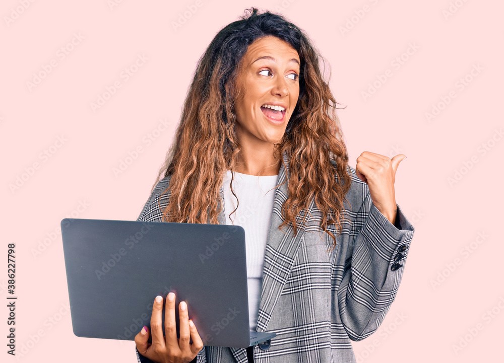 Young hispanic woman with tattoo holding laptop pointing thumb up to the side smiling happy with open mouth Young hispanic woman with tattoo holding laptop pointing thumb up to the side smiling happy with open mouth