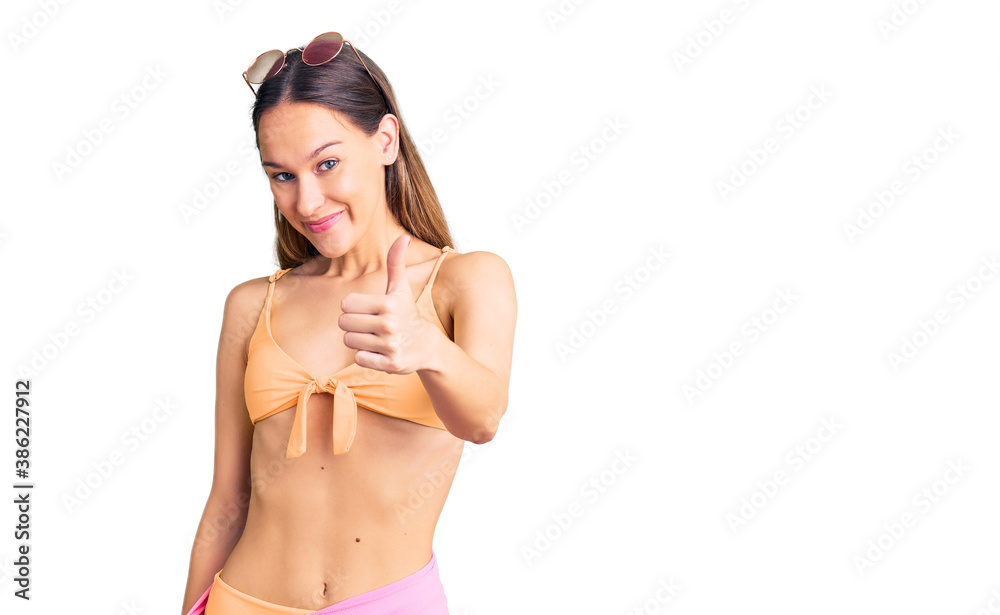 Beautiful brunette young woman wearing bikini and sunglasses doing happy thumbs up gesture with hand. approving expression looking at the camera showing success.