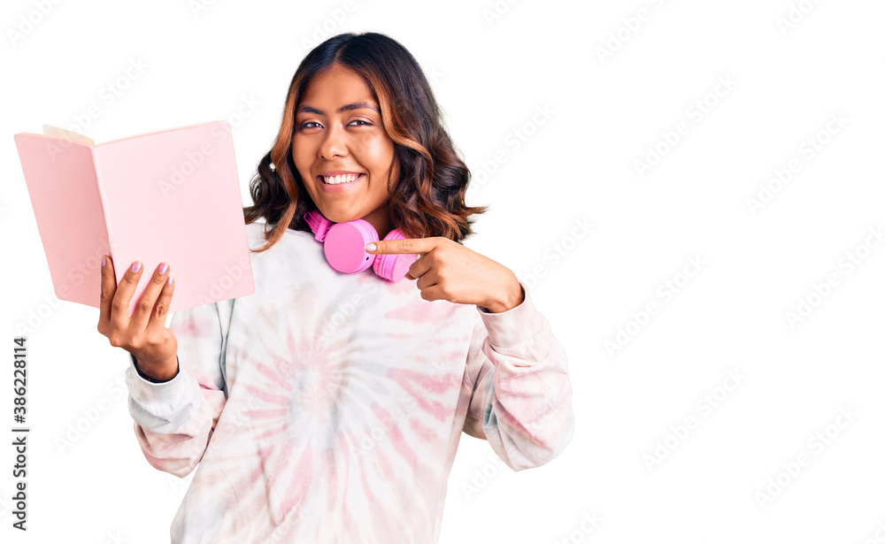Young beautiful mixed race woman reading a book wearing headphones smiling happy pointing with hand and finger Young beautiful mixed race woman reading a book wearing headphones smiling happy pointing with hand and finger