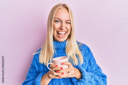 Quadro em tela Young blonde girl wearing winter sweater and drinking a cup of hot coffee winking looking at the camera with sexy expression, cheerful and happy face