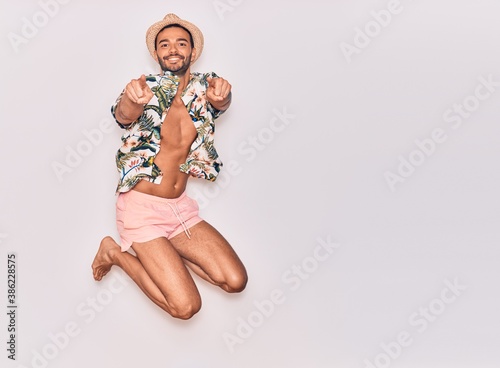 Young handsome hispanic man on vacation wearing swimwear, floral shirt and hat smiling happy. Jumping with smile on face pointing with fingers over isolated white background