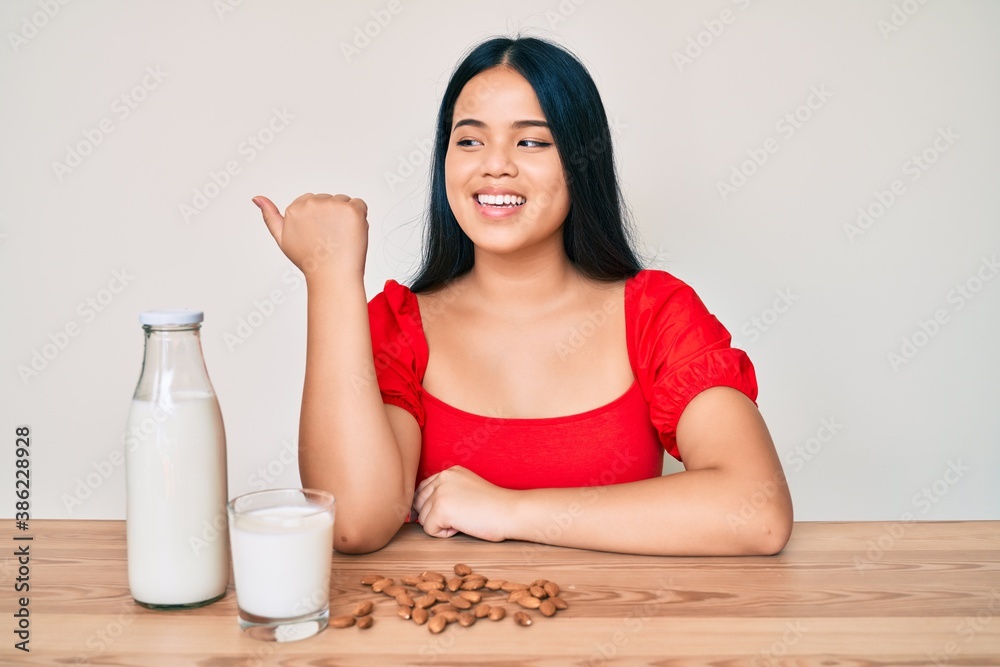 Young asian girl drinking healthy almond milk pointing thumb up to the side smiling happy with open mouth Young asian girl drinking healthy almond milk pointing thumb up to the side smiling happy with open mouth