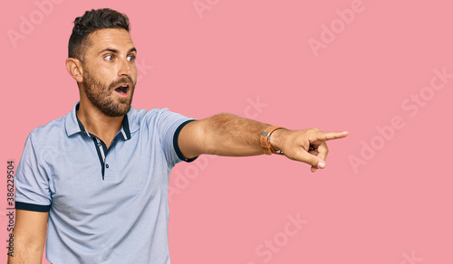 Handsome man with beard wearing casual clothes pointing with finger surprised ahead, open mouth amazed expression, something on the front