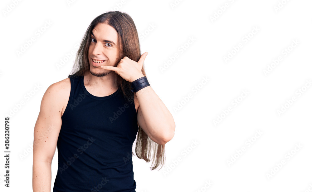 Young adult man with long hair wearing goth style with black clothes smiling doing phone gesture with hand and fingers like talking on the telephone. communicating concepts. Young adult man with long hair wearing goth style with black clothes smiling doing phone gesture with hand and fingers like talking on the telephone. communicating concepts.